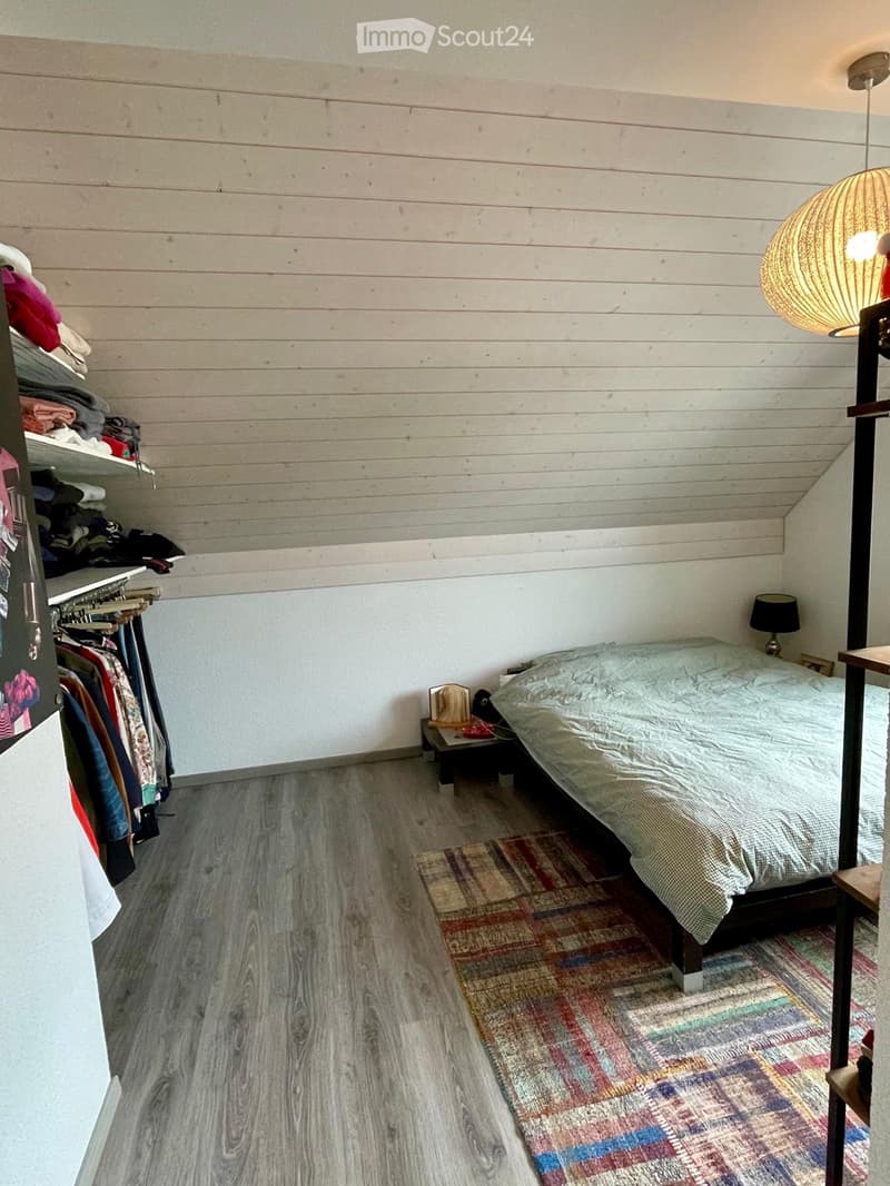 Cozy bedroom with wooden walls, a double bed with a patterned duvet, a nightstand with a lamp, a colorful rug, and a wardrobe with hanging clothes.
