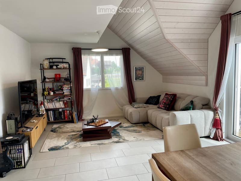Living room with a sectional sofa, coffee table, bookshelf, dining table, and a guitar. The room features large windows with curtains and a slanted wooden ceiling.