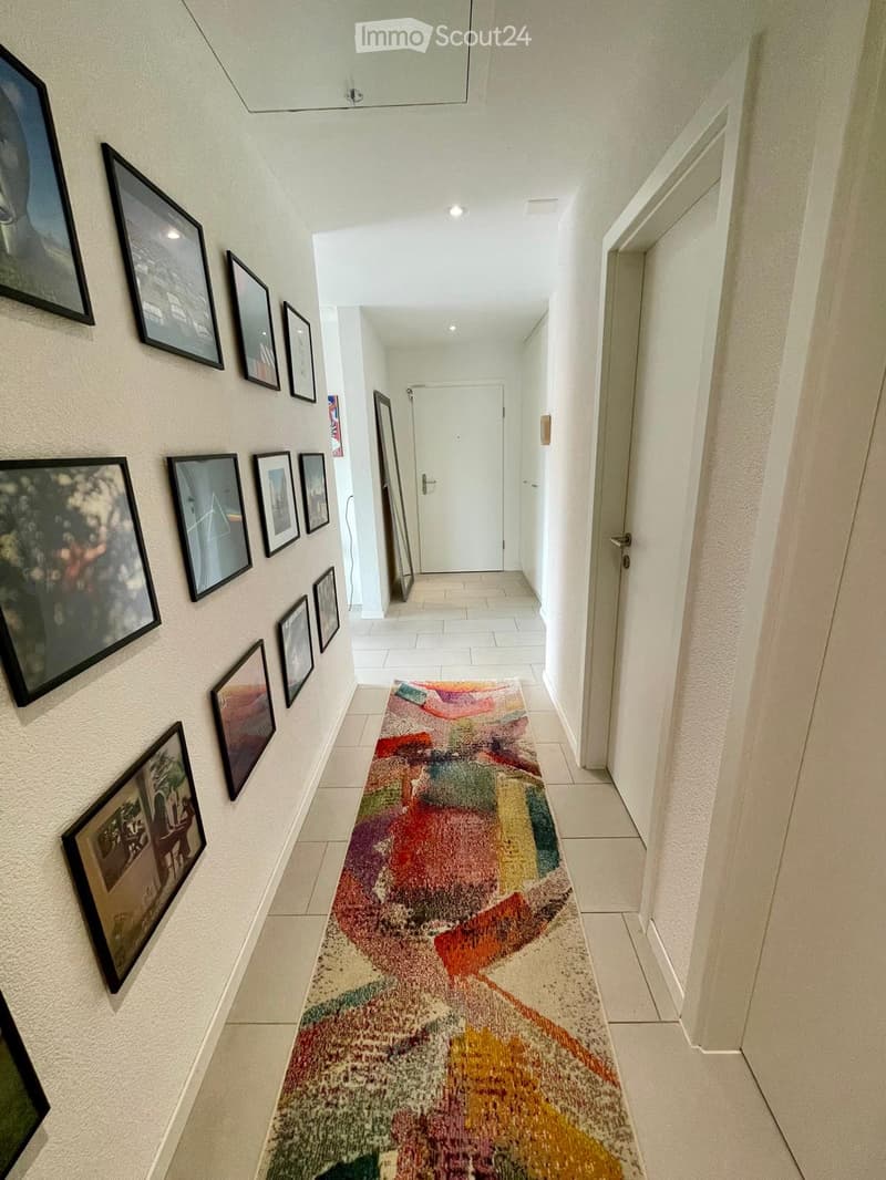 Hallway with white walls, tiled floor, colorful runner rug, and framed artwork displayed on one wall. A white door is visible at the end of the hallway.
