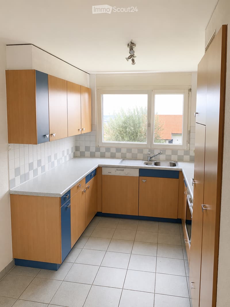 Modern kitchen with light wooden cabinets, white countertops, and a stainless steel sink. The space features tiled flooring and a window offering a view of greenery outside.