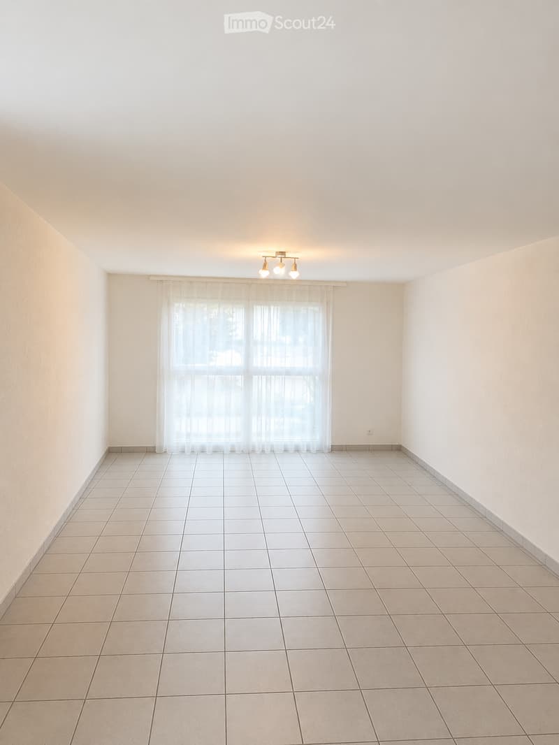 Empty room with tiled floor, white walls, large window with sheer curtains, and a ceiling light fixture.