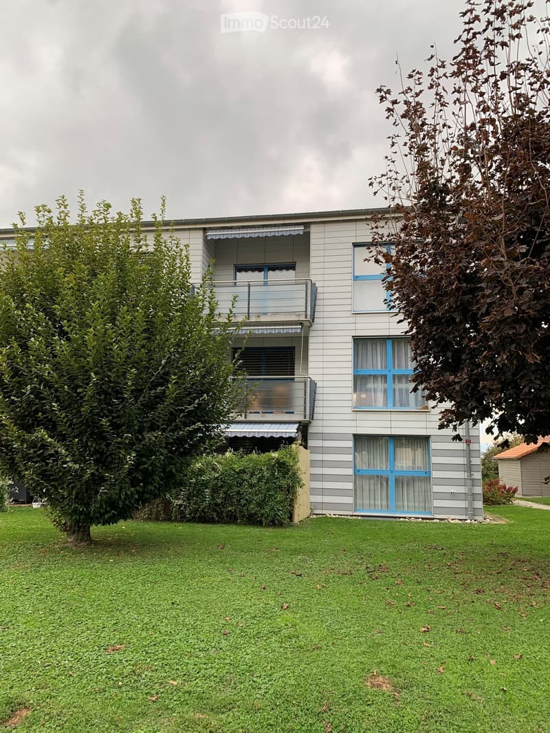 Two-story house with grey siding, blue-framed windows, and balconies. The house is surrounded by a well-maintained lawn and trees.