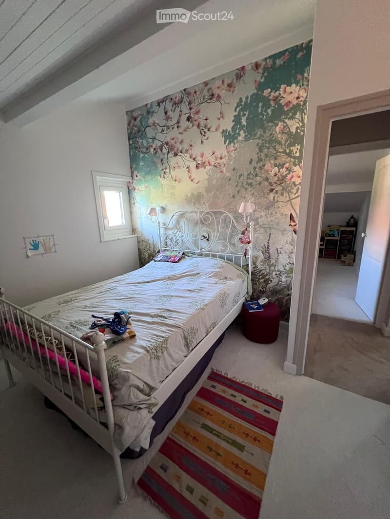 Cozy bedroom with a white metal bed, colorful rug, and floral wallpaper. Includes a small round stool and table lamp.