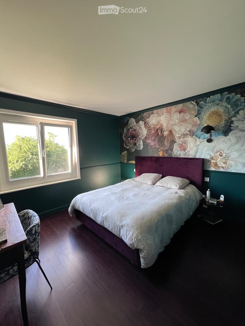 Bedroom with a large bed, nightstand, chair, and table. The walls are painted in dark green with a floral mural above the bed. The room has wooden flooring and large windows letting in natural light.