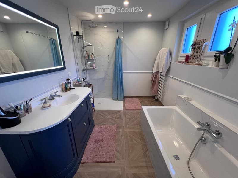 Modern bathroom with a double sink vanity, bathtub, large mirror, and shower area. Features wooden flooring, white walls, and decorative towels.