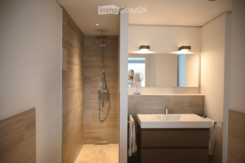 Modern bathroom with a large mirror, a single sink with a modern faucet, a shower area with a handheld showerhead, and a vanity cabinet. The walls are tiled in a light wood finish, and the floor is also wood-tiled.