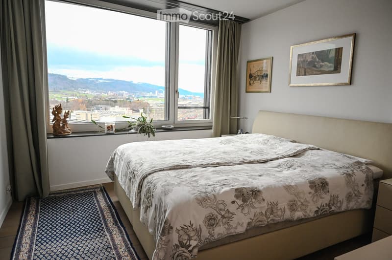 A well-lit bedroom with a large window offering a scenic view, featuring a neatly made bed with a floral duvet, a nightstand, a small plant on the windowsill, and decorative picture frames on the wall.