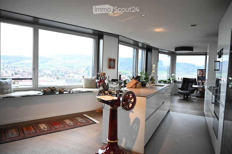 Modern kitchen with large windows offering a scenic view, featuring a kitchen island, cabinets, a sink, a stool, a chair, and a framed picture on the wall.