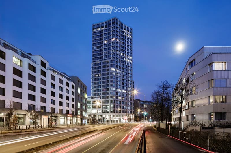 Modern high-rise building with numerous windows, surrounded by other contemporary structures and illuminated streetlights.