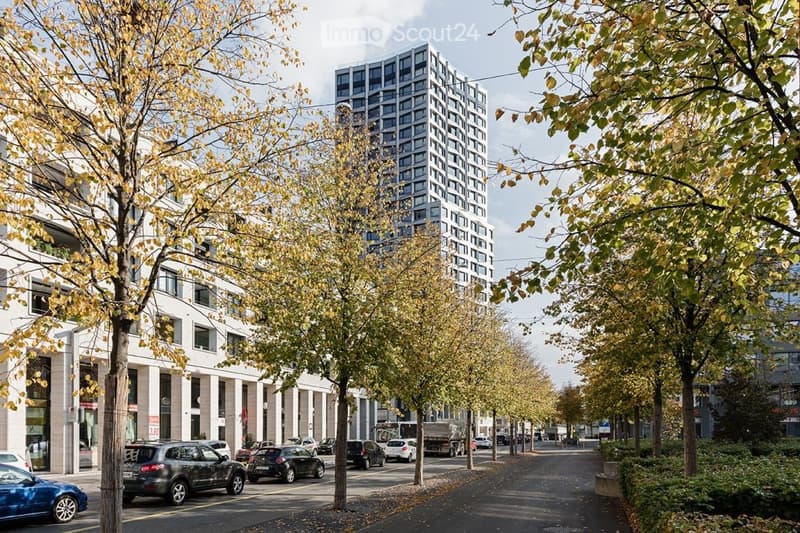 Urban street lined with trees, featuring modern multi-story buildings, parked cars, and a clear sky.