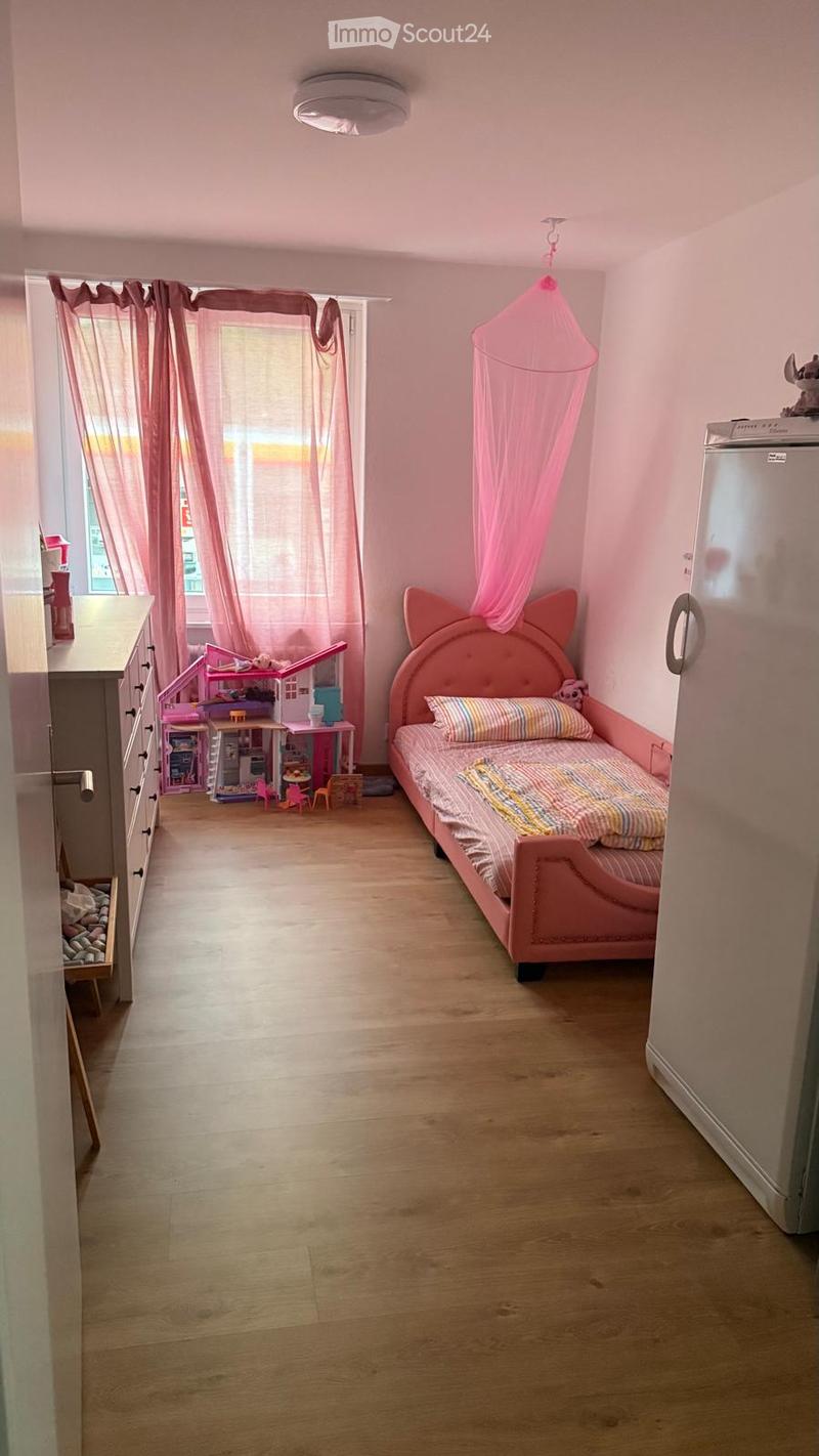 A child’s bedroom with a pink bed, a toy house, a dresser, and a mosquito net hanging from the ceiling. The room has wooden flooring and large windows with pink curtains.