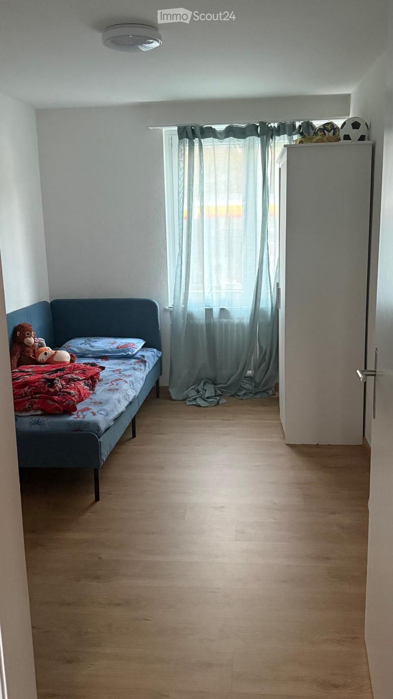 A small bedroom with a single bed, pillows, a blanket, toys, and a wardrobe. The room has wooden flooring and a window with curtains.