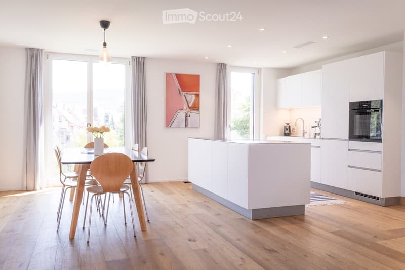 Modern kitchen and dining area with white cabinets, a central island, a dining table with chairs, and a vase of flowers on the table. The space features wooden flooring and large windows with curtains.