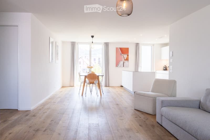 Bright, open-plan living and dining area with light wood flooring, white walls, and large windows. Features a grey sofa, dining table with chairs, and a small kitchen area with cabinets.