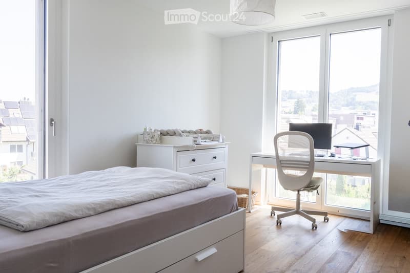 Modern bedroom with a large bed, a desk with a computer and chair, a drawer unit, and a wicker basket. Large windows offer a scenic view.