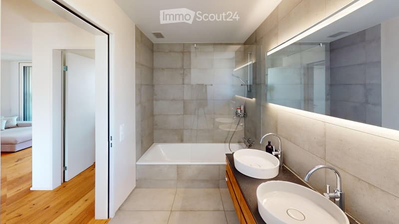 Modern bathroom with a bathtub, double sink, large mirror, and wooden cabinet. The space features light-colored tiles and a glass-enclosed shower area.