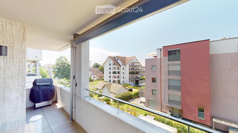 Balcony with tiled floor, metal railing, grill, and a view of surrounding buildings and greenery.