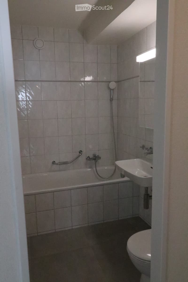 Bathroom with a bathtub, toilet, sink, and a handrail for accessibility. The walls are tiled with grey tiles, and the floor is dark grey.