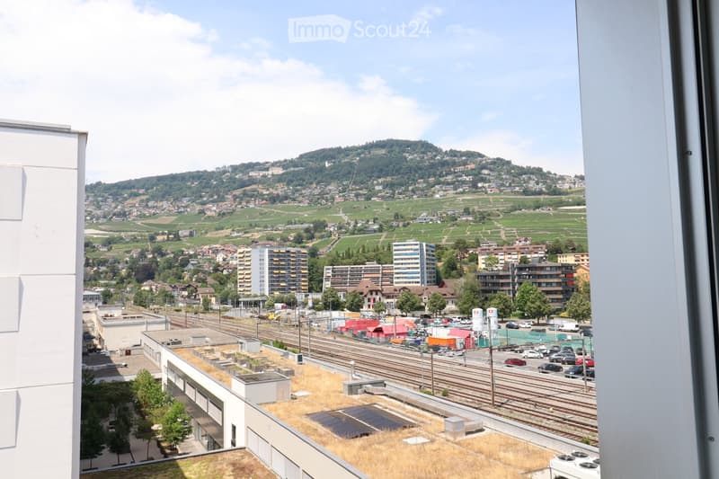 View from a balcony showing a cityscape with buildings, train tracks, and a distant hillside with vineyards and houses.