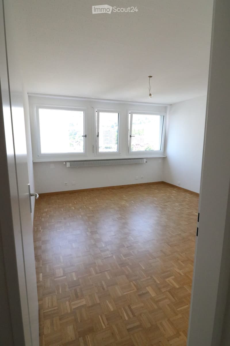 Empty room with wooden herringbone flooring, white walls, and three windows with white frames. The room has a simple ceiling light fixture and a radiator below the windows.