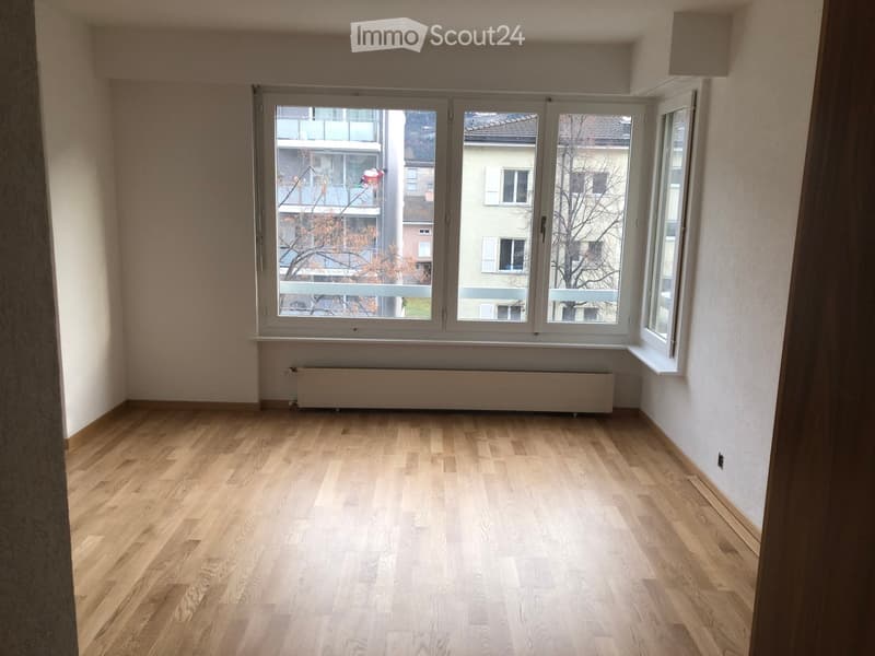Empty room with wooden flooring, large windows, radiator, and view of neighboring buildings.