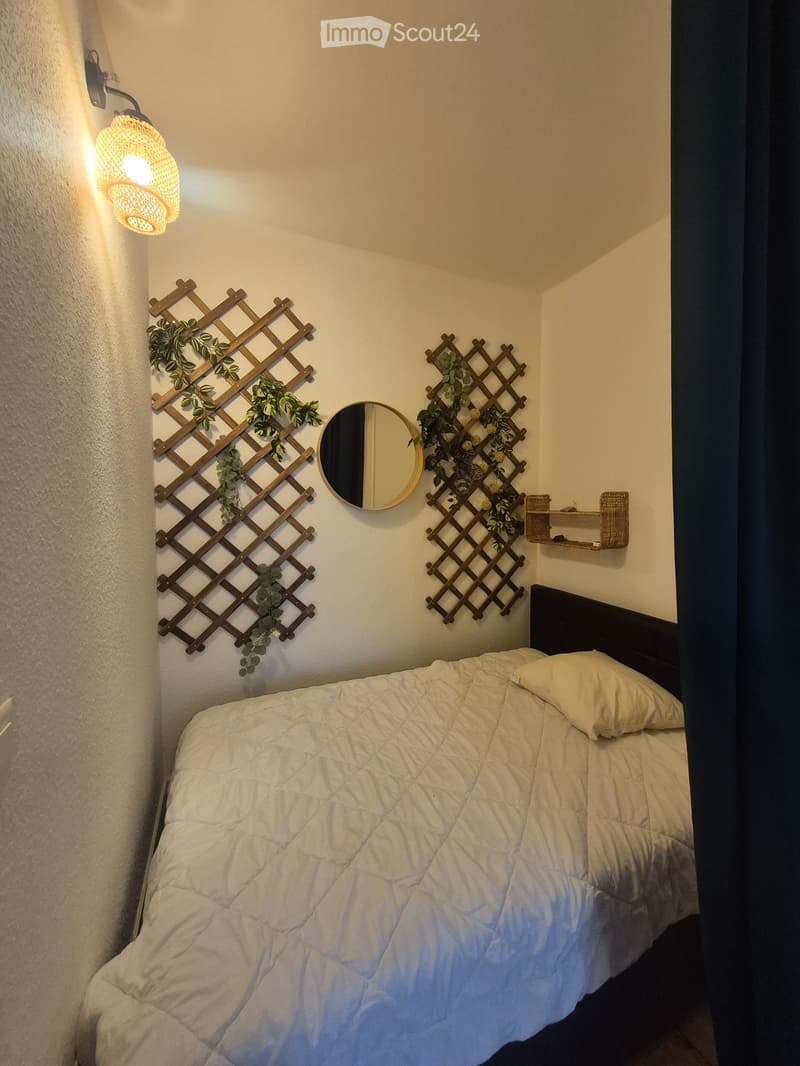 Cozy bedroom with a neatly made bed, a round wall mirror, decorative wooden lattice panels with greenery, and a small shelf with a woven basket.