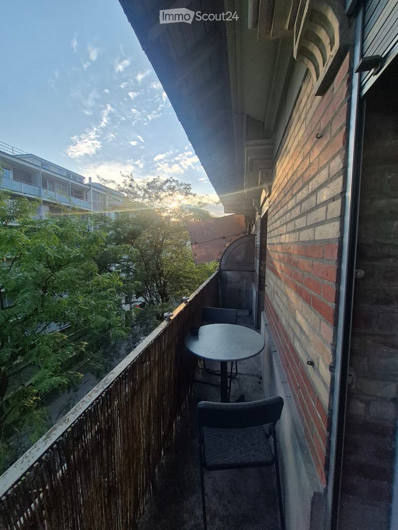 Balcony with a round table and two chairs, overlooking a tree-lined area with neighboring buildings.