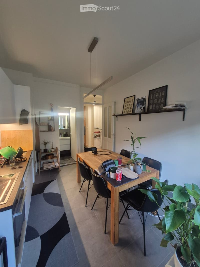 A modern dining area with a wooden table, black chairs, and various items including cups, a bottle, and a plant. The room features a sleek design with a visible kitchen area in the background.