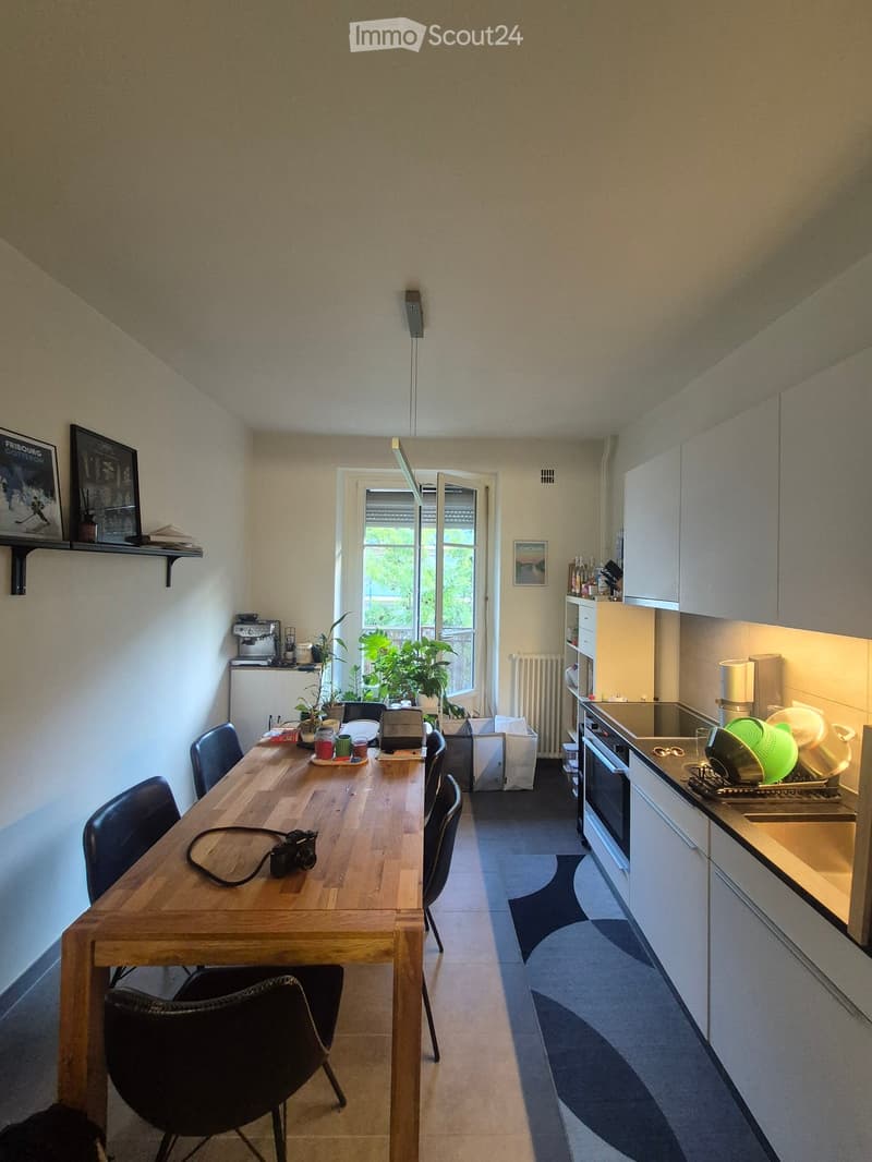 Modern kitchen with a wooden dining table, chairs, and various appliances including an oven, sink, and fridge. There are plants, a camera, and a framed picture on the wall.