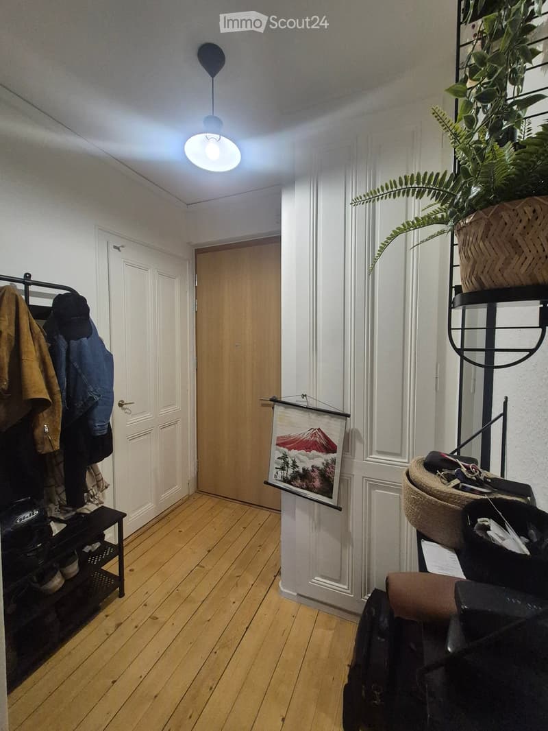 A narrow hallway with wooden flooring, white walls, and a pendant light fixture. There are two doors, a coat rack with jackets, a small shelf with shoes, and a potted plant on a stand.
