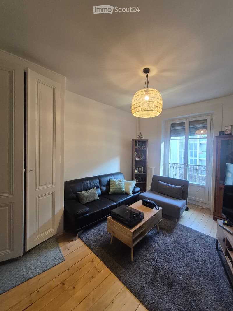 Modern living room with wooden flooring, a black leather sofa, a grey armchair, a wooden coffee table, a hanging woven lamp, a shelf, and a television.