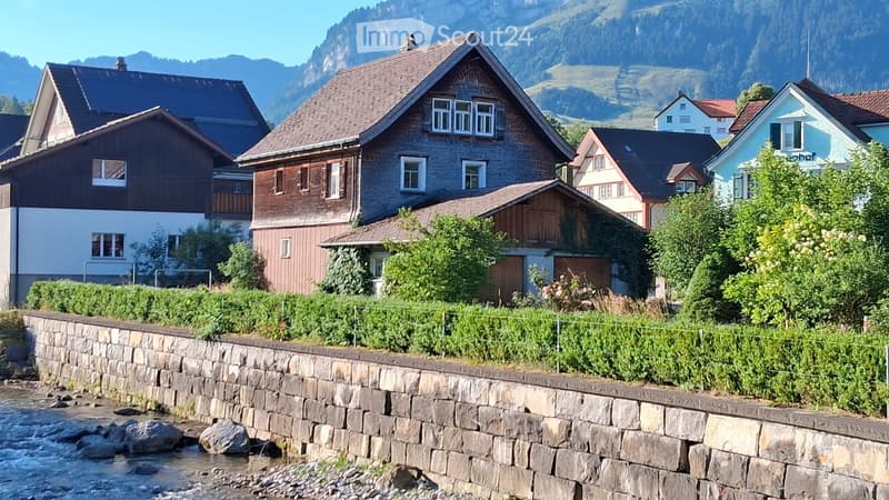 A row of traditional houses with pitched roofs and wooden facades, surrounded by greenery and a stone wall along a river.