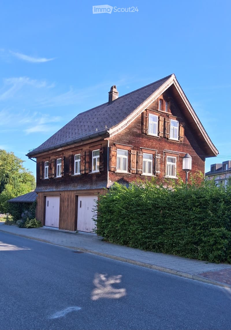 Two-story wooden house with a steeply pitched roof, multiple windows with shutters, and a garage with two doors.