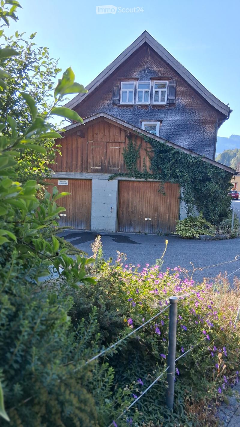 Two-story house with a gabled roof, brick facade, and wooden garage doors. Ivy climbs up the side of the house, and there is a garden with purple flowers in the foreground.