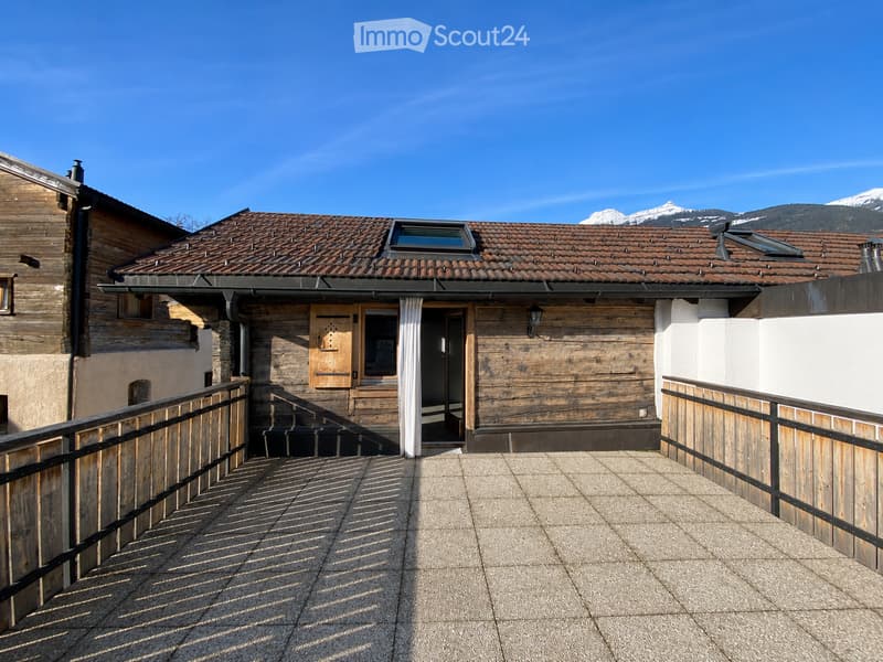 A wooden building with a tiled roof, featuring a balcony area with a wooden railing and a view of mountains in the background.