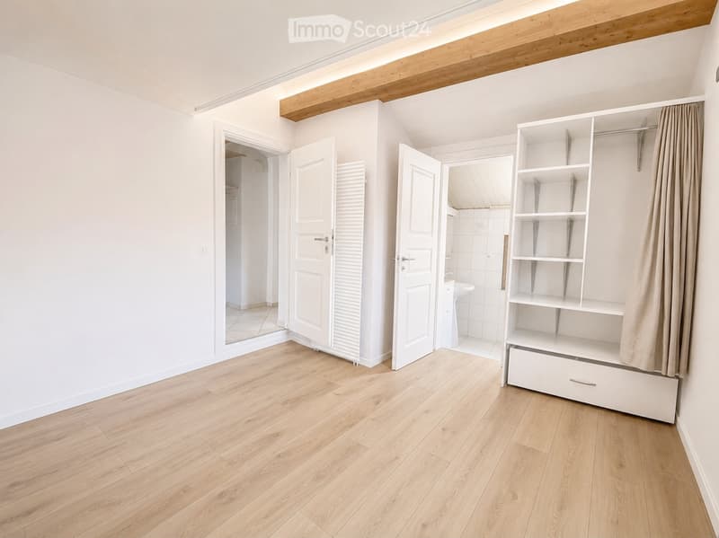 Empty room with light wooden flooring, white walls, and built-in shelving unit. Open doors lead to adjacent spaces, including a bathroom.