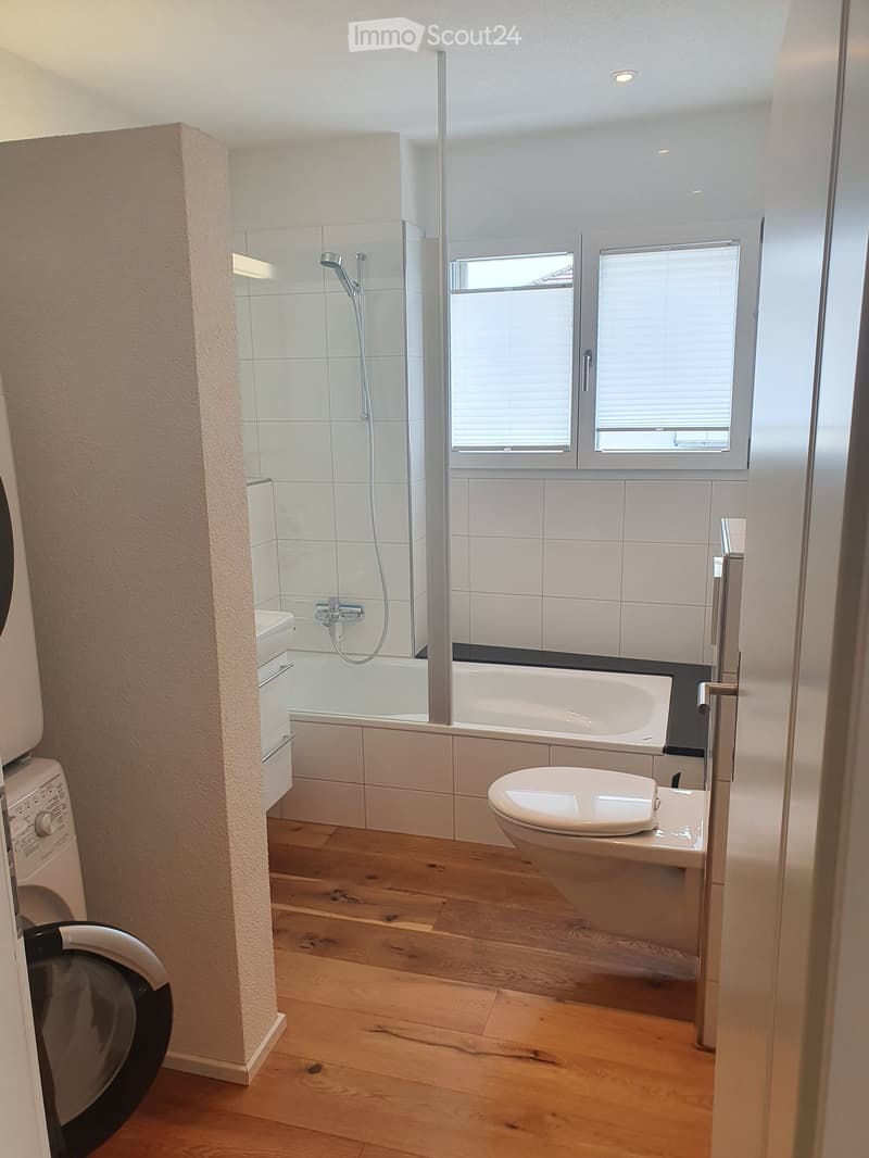 Modern bathroom with wooden flooring, white tiled shower area, bathtub, toilet, and washing machine visible on the left side.
