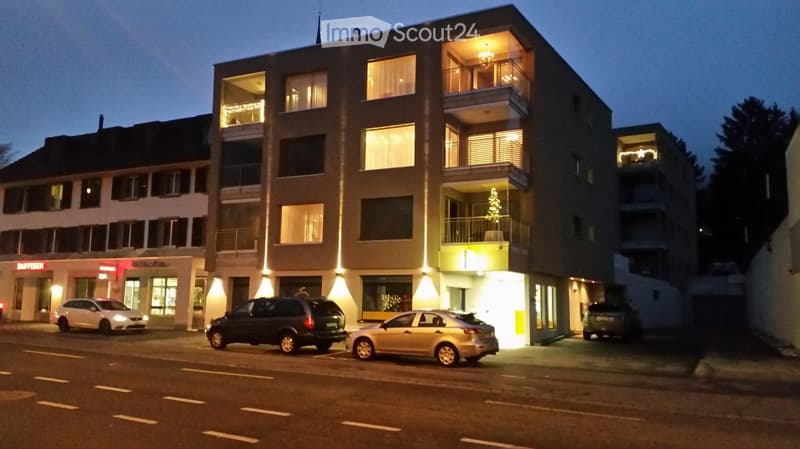 Multi-story apartment building with illuminated windows and balconies, several cars parked in front.