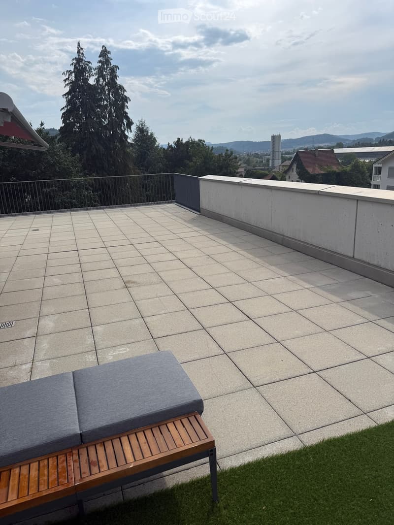 Rooftop terrace with tiled floor, low concrete wall, metal railing, and a wooden bench with a grey cushion.