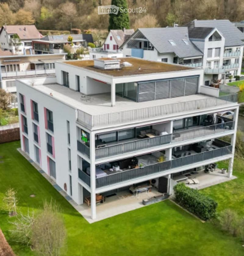 Modern multi-story house with white exterior, grey roof, multiple balconies, and a green lawn in front.
