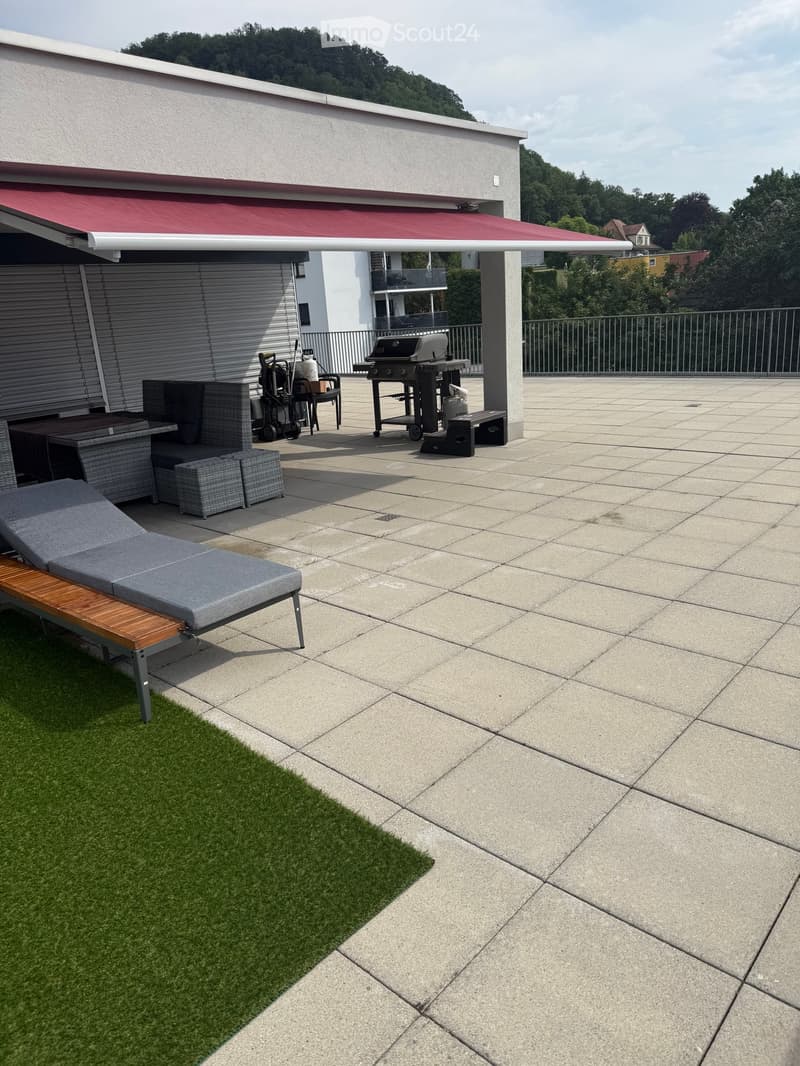 A modern outdoor patio with a sun lounger, a small seating area, a grill, and a storage box, featuring a tiled floor and a red awning.