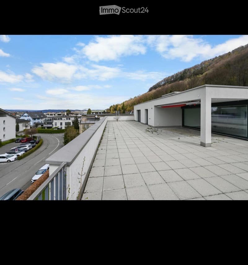 Balcony with tiled floor, low wall, and modern building with garage in the background. The view includes a road with parked cars and a hillside with trees.