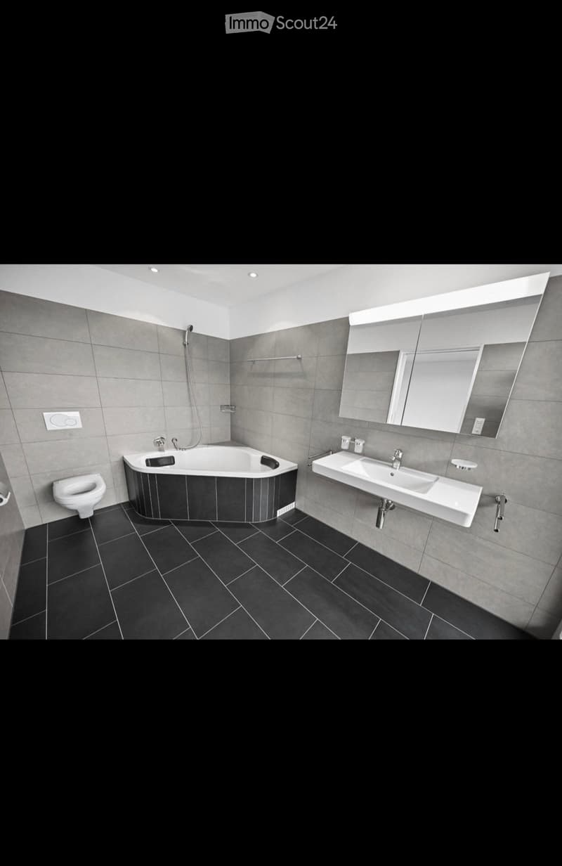 Modern bathroom with dark tiled floor, grey walls, bathtub, toilet, double sink, large mirror, and modern lighting.