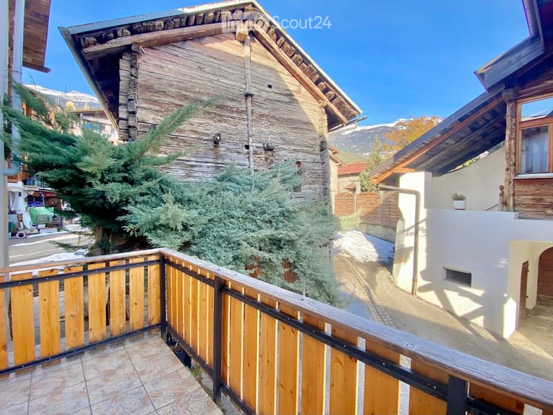 Balcony with wooden railing, stone-tiled floor, overlooking a stone house with wooden accents and surrounding greenery.