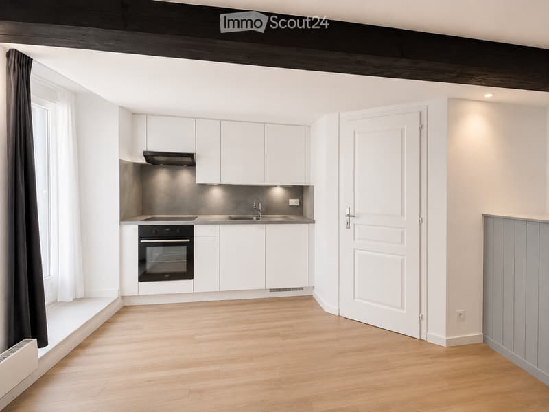 Empty room with wooden flooring, white cabinetry, and a modern kitchen area featuring a sink, stove, and oven. The space is well-lit with large windows and a door leading to another room.