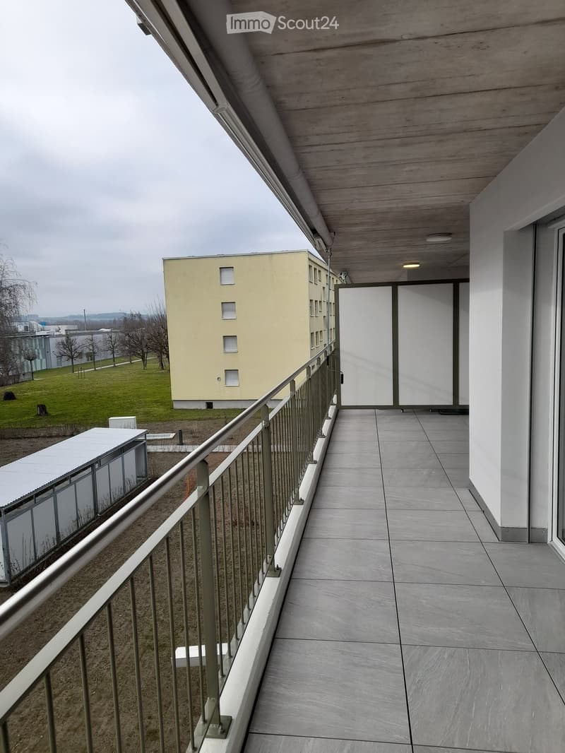Modern balcony with tiled floor, metal railing, and access to a building entrance. The view includes a grassy area, a small structure, and a multi-story building in the background.