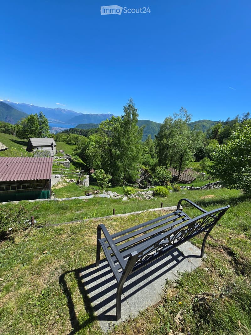 A metal bench sits on a concrete platform overlooking a lush green valley with scattered houses and distant mountains.