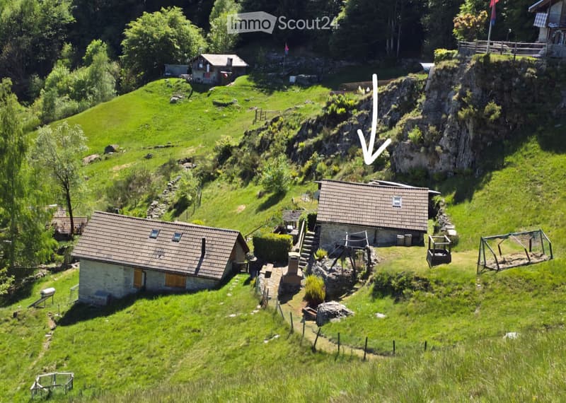 A scenic outdoor area featuring several small houses and structures nestled in a lush, green landscape. The area includes stone and wooden houses, a raised wooden platform, and a rocky cliffside. The terrain is grassy with scattered trees and shrubs, crea