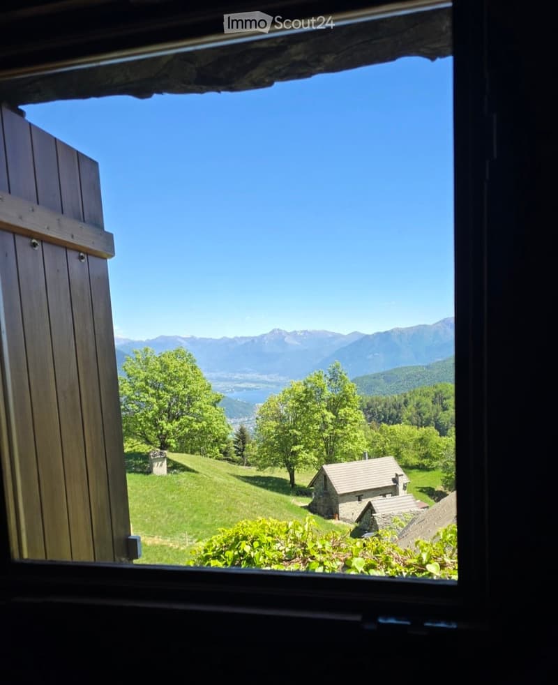 View from a window showing a lush green landscape with trees, a small stone house, and distant mountains under a clear blue sky.