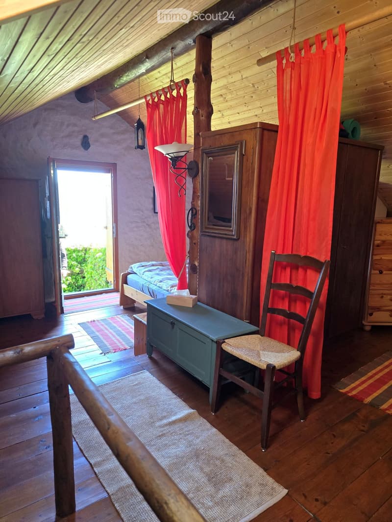 Cozy attic bedroom with wooden walls and slanted ceilings, featuring a bed, a wooden chair, a small table, a mirror, and red curtains.